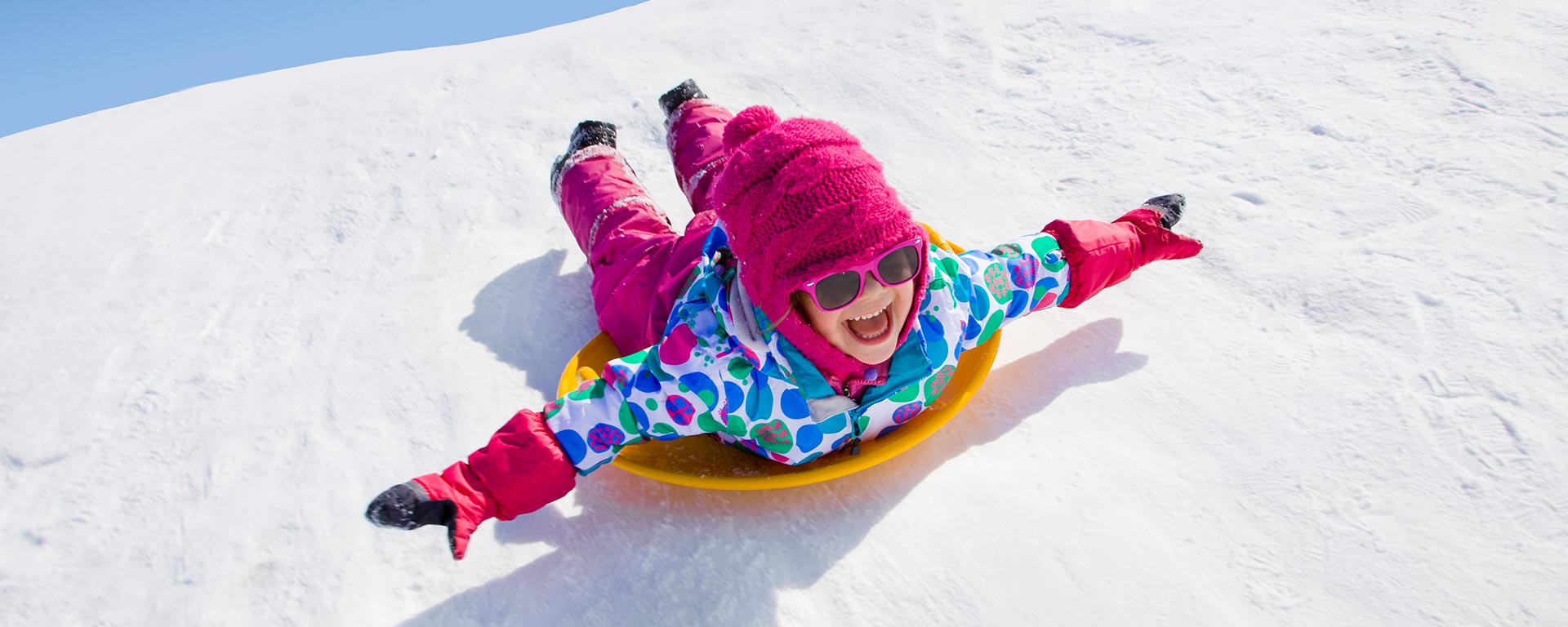 a child dressed in pink and bluish is riding a plastic snow sled laying on it's stomach